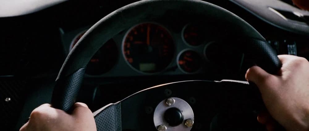 La imagen muestra un primer plano de una persona sosteniendo el volante de un coche. En el fondo, se puede ver el tablero con varios indicadores, incluyendo un velocímetro y un tacómetro. Esta imagen es interesante y relevante ya que captura la perspectiva de un conductor, enfatizando el control y la concentración necesarios al conducir.