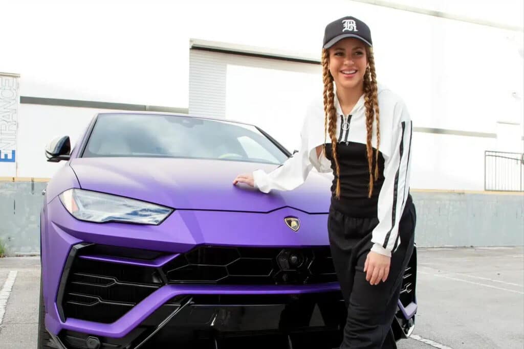 La imagen muestra a Shakira de pie junto a un Lamborghini Urus morado. Shakira lleva un atuendo en blanco y negro y una gorra negra. La imagen sugiere que se está promocionando un sorteo o concurso en el que se puede ganar un Lamborghini.