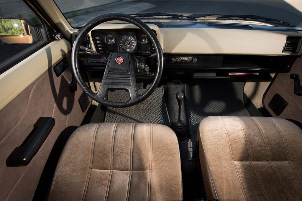 La imagen muestra el interior de un Talbot Horizon. El área del asiento del conductor y el tablero son claramente visibles. El volante está en el centro, con un logotipo en el medio. El tablero incluye varios indicadores y controles, como un velocímetro y un indicador de combustible. La palanca de cambios está ubicada entre los dos asientos delanteros. Los asientos están tapizados en tela marrón, y los paneles de las puertas también son visibles, con manijas y manivelas para las ventanas. El diseño general parece ser de un vehículo de modelo antiguo, posiblemente de finales del siglo XX. Esta imagen es interesante porque proporciona una visión del diseño y la disposición de los interiores de coches de una era pasada, destacando la simplicidad y funcionalidad de los controles y características.