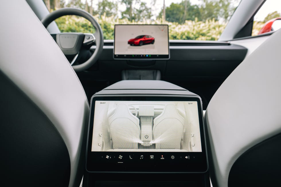 Interior de un Tesla Model 3 con diseño minimalista y moderno. La imagen muestra dos pantallas táctiles: una central en la consola delantera que exhibe la imagen del coche, y otra en la parte trasera del reposabrazos central, enfocada en los controles del asiento. Los asientos tienen un acabado en cuero blanco, destacando el contraste con los elementos oscuros del salpicadero. El fondo incluye un paisaje verde visible a través del parabrisas.