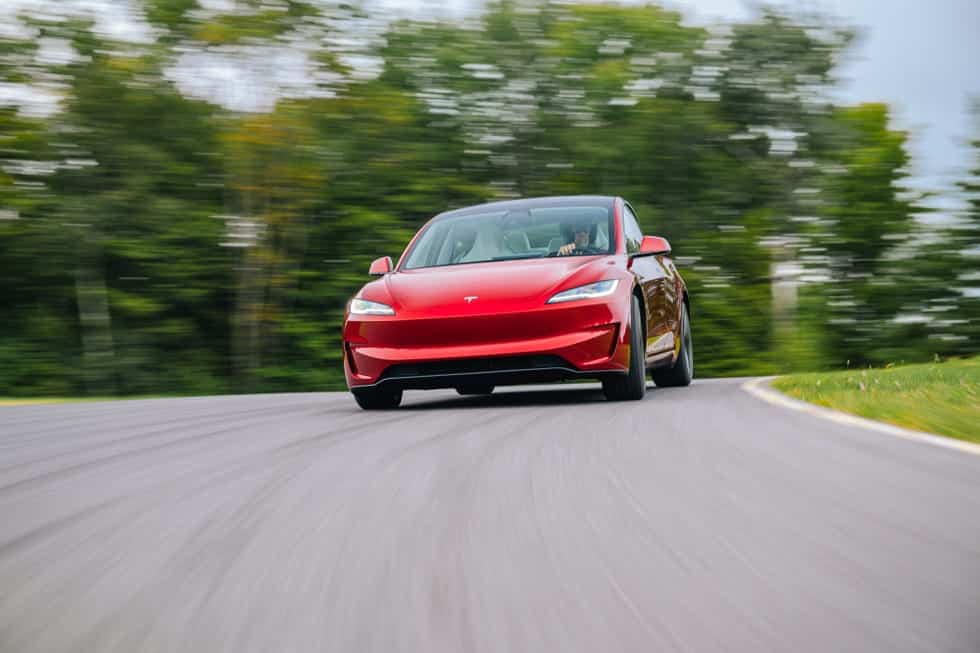 La imagen muestra un coche Tesla rojo conduciendo en una carretera curva con un fondo borroso de árboles, lo que indica movimiento y velocidad. El coche está en el centro de la imagen, destacando su diseño frontal. Esta imagen es interesante y relevante ya que muestra el diseño elegante y el rendimiento del vehículo Tesla, destacando su capacidad para manejar curvas y velocidad de manera eficiente.