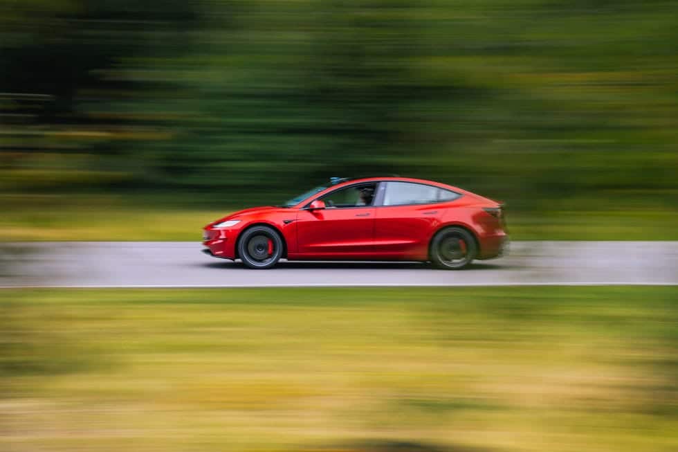 Un Tesla Model 3 rojo, de perfil, avanzando a gran velocidad por una carretera. El coche tiene un diseño aerodinámico con líneas elegantes, techo de cristal oscuro y llantas negras deportivas. El fondo muestra vegetación desenfocada, lo que resalta la sensación de movimiento.