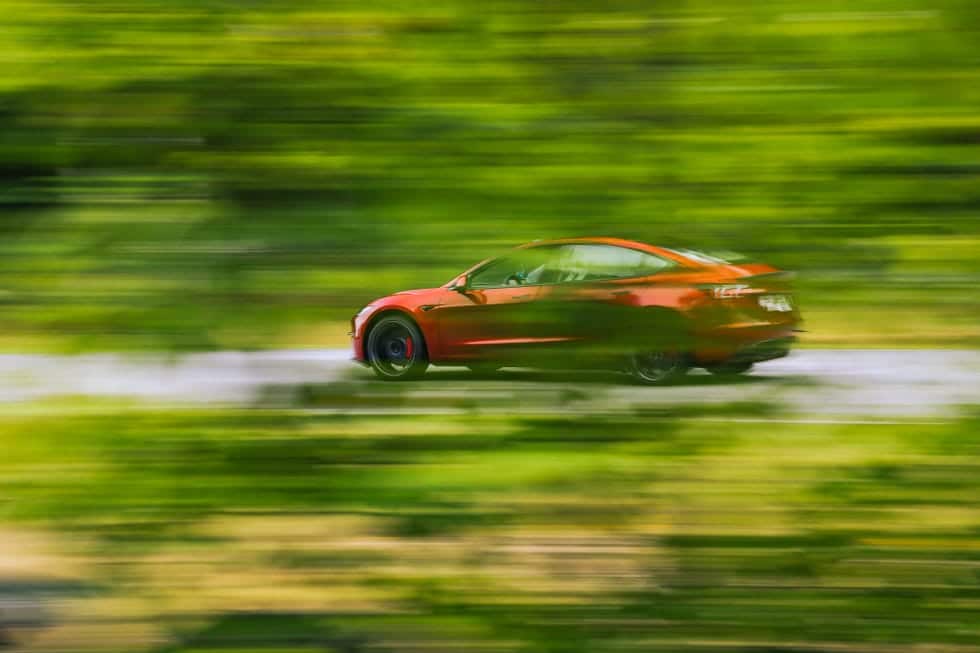 Un Tesla Model 3 rojo, visto de perfil mientras avanza a gran velocidad por una carretera rodeada de un entorno verde muy desenfocado, lo que acentúa el dinamismo de la imagen. El coche muestra su diseño moderno y aerodinámico, con líneas suaves, techo de cristal y llantas negras deportivas.