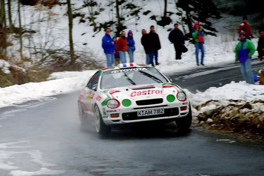 La imagen muestra un coche de rally, específicamente un Toyota Celica GT-Four, participando en un evento de rally en una carretera nevada y helada. El coche está adornado con calcomanías de patrocinio de Castrol y tiene la matrícula "K-AM 7182". La escena está ambientada en un entorno invernal con nieve en el suelo y espectadores observando desde el borde de la carretera. El coche parece estar navegando una curva, lo que resalta la habilidad y precisión necesarias en la conducción de rally, especialmente bajo condiciones climáticas desafiantes. ¿Podría el nuevo Toyota Celica recuperar esto?