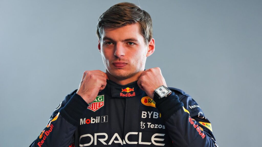 "La imagen muestra a Max Verstappen, piloto de Fórmula 1, con su traje de carreras de Red Bull Racing. Está sujetando el cuello del traje con ambas manos, y se pueden ver los logotipos de los patrocinadores, incluyendo Red Bull, Oracle, Mobil 1, Tezos y Tag Heuer. El rostro de Verstappen está desenfocado, pero es reconocible por su postura y el contexto.