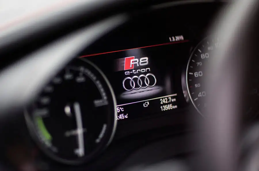 Claro, aquí tienes: La imagen muestra el tablero de instrumentos de un Audi R8 e-tron. La pantalla central muestra de manera prominente el logotipo "R8 e-tron" junto con los anillos de Audi. El medidor de la izquierda parece ser un medidor de energía, mientras que el de la derecha es un velocímetro. La pantalla también muestra información adicional, incluyendo la temperatura (15°C), la hora (5:45), la distancia recorrida (242.7 km) y el kilometraje total (13566 km). Esta imagen es interesante porque destaca la interfaz digital y la marca de un vehículo eléctrico de alto rendimiento.