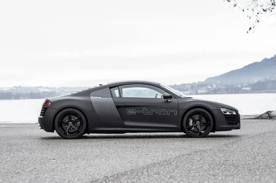 Claro, aquí tienes: La imagen muestra un elegante coche deportivo negro con la palabra "e-tron" destacada en su lateral. El coche está estacionado sobre una superficie pavimentada cerca de un cuerpo de agua, con un paisaje montañoso al fondo. La marca "e-tron" indica que se trata de un vehículo eléctrico, lo cual es interesante y relevante debido a la creciente tendencia e importancia de los coches eléctricos en la industria automotriz para la sostenibilidad ambiental.