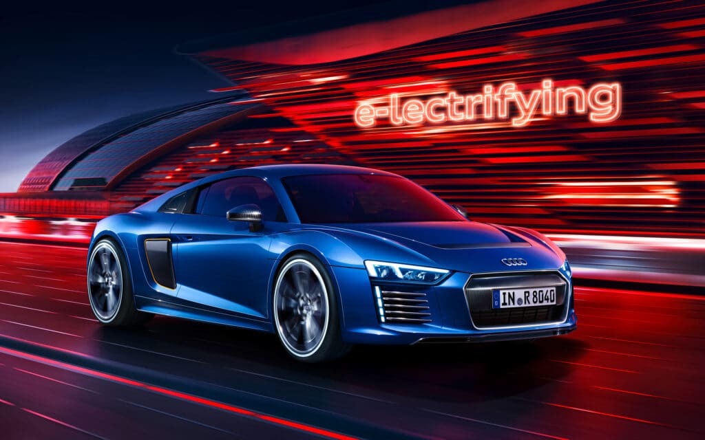 La imagen muestra un Audi R8 e-tron azul conduciendo a alta velocidad con un fondo futurista que presenta rayos de luz rojos y naranjas. El fondo incluye un edificio moderno y el texto "e-lectrifying" en estilo neón, enfatizando la naturaleza eléctrica del vehículo. El diseño del coche es elegante y aerodinámico, con distintivos faros LED y una parrilla frontal prominente. La matrícula dice "IN R 8040". Esta imagen es interesante y relevante ya que destaca la tecnología avanzada y el rendimiento del Audi R8 e-tron, mostrando su atractivo de manera visualmente impactante.