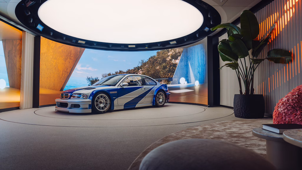 La imagen muestra la vista trasera de un BMW M3 GTR, un coche de carreras de alto rendimiento, estacionado en un garaje o taller con poca iluminación. El coche cuenta con un gran alerón trasero, lo que indica que está diseñado para carreras de alto rendimiento. La iluminación en el garaje resalta las características aerodinámicas del coche y su diseño elegante, haciéndolo visualmente impactante y relevante para los entusiastas del automovilismo o aquellos interesados en la ingeniería de coches de carreras. Este coche es famoso por su aparición en el videojuego 'Need for Speed: Most Wanted', donde juega un papel central.