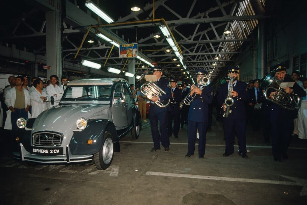 La imagen muestra un grupo de personas reunidas alrededor de un Citroën 2CV clásico dentro de lo que parece ser una instalación industrial o de fabricación. Hay una banda tocando instrumentos musicales, incluyendo tubas y trompetas, lo que sugiere un evento de celebración. El coche tiene un cartel en la parte delantera que dice "DERNIÈRE 2 CV", indicando que este podría ser el último modelo del Citroën 2CV que se produce. Esta imagen es interesante y relevante ya que captura un momento significativo en la historia automotriz, marcando el final de la producción de un vehículo icónico.