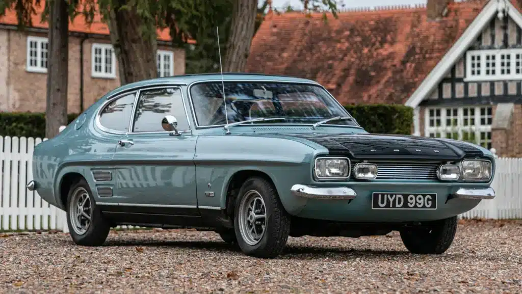 La imagen muestra un Ford Capri Mk1 de 1969. El coche está estacionado sobre una superficie de grava, con una casa de fondo que tiene una cerca blanca y árboles alrededor. El coche tiene una carrocería azul claro con un capó negro, parachoques cromados y faros redondos distintivos. La matrícula dice "UYD 99G". Esta imagen es interesante y relevante porque muestra un vehículo vintage que es icónico en la historia automotriz, conocido por su diseño elegante y rendimiento en su época.