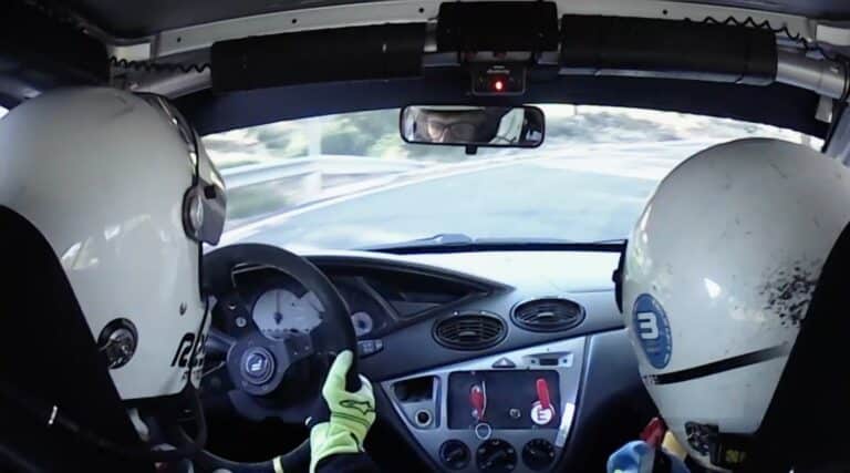La imagen muestra el interior de un coche de rally con dos personas usando cascos y equipo de carreras. La persona a la izquierda está conduciendo el coche, sujetando el volante, mientras que la persona a la derecha parece ser el copiloto o navegante. El salpicadero y el panel de control del coche son visibles, con varios interruptores e indicadores. La vista a través del parabrisas muestra una carretera sinuosa, indicando que el coche está en movimiento, probablemente durante una carrera de rally. Esta imagen es interesante y relevante ya que captura el entorno dinámico e intenso de las carreras de rally, destacando el trabajo en equipo y la concentración requeridos tanto por el conductor como por el copiloto.