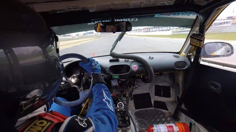 La imagen muestra el interior de un coche de carreras desde la perspectiva del conductor. El conductor lleva un casco y un traje de carreras, y está sujetando el volante con ambas manos. El salpicadero es visible, con varios controles y una pantalla digital. Hay un extintor montado en el suelo del coche. El coche está en una pista de carreras, y la pista es visible a través del parabrisas. Esta imagen es interesante y relevante porque proporciona una vista interna de la experiencia de conducir un coche de carreras, destacando la perspectiva del conductor y la configuración del interior del coche.