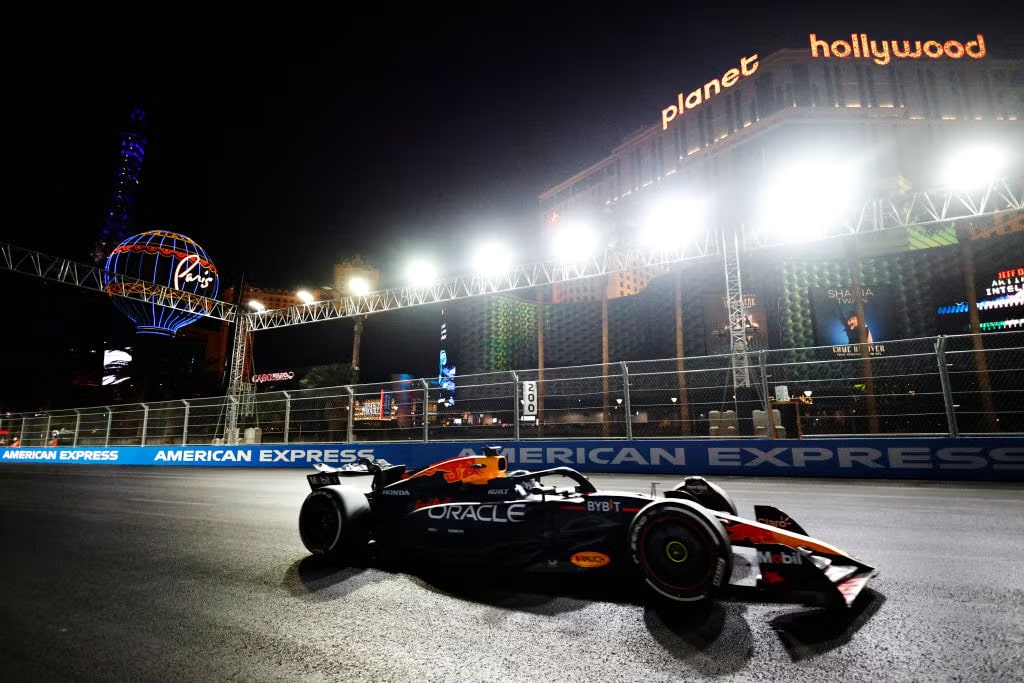 La imagen muestra un coche de Fórmula 1 de Red Bull Racing, conducido por Max Verstappen, en un circuito urbano nocturno. El coche está decorado con los logotipos de patrocinadores como Oracle, Bybit y Mobil. En el fondo, se pueden ver los letreros iluminados de "Planet Hollywood" y "Paris" en Las Vegas, lo que indica que este evento se lleva a cabo en el Strip de Las Vegas. La pista está bordeada por barreras y anuncios de American Express. La escena es vibrante y dinámica, capturando la emoción de un evento de automovilismo de alto perfil en una ubicación famosa.