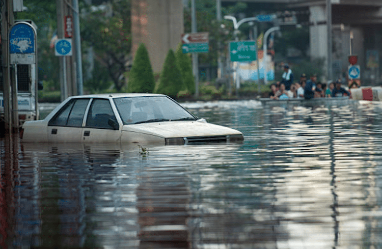 La imagen muestra una calle inundada con un coche blanco sumergido hasta las ventanas en el agua. En el fondo, hay personas caminando por el agua y señales de tráfico parcialmente cubiertas por la inundación.