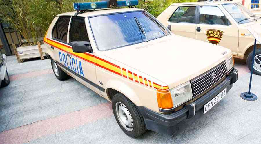 La imagen muestra un coche clásico de policía con la palabra "POLICIA" escrita en letras azules en el costado. El coche es de color beige con franjas rojas y amarillas a lo largo del lateral. En el techo del coche hay una barra de luces azules. La matrícula dice "CPN 2210 A". En el fondo se puede ver otro vehículo similar con las palabras "POLICIA NACIONAL" en la puerta. El entorno parece ser una zona exterior, posiblemente una exhibición o museo. Esta imagen es interesante porque muestra un modelo antiguo de vehículo policial, que puede tener un significado histórico o ser de interés para quienes estudian la historia de las fuerzas del orden.