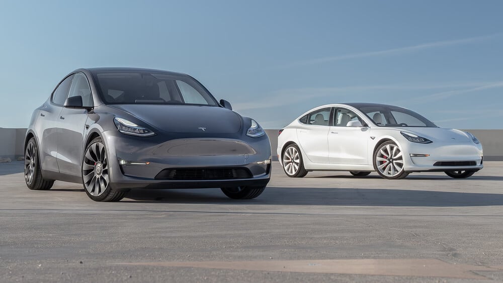 Imagen de un Tesla Model Y de color oscuro y un Tesla Model 3 blanco estacionados sobre una superficie de concreto bajo un cielo despejado. Ambos coches están posicionados en ángulo, mostrando sus perfiles frontales y laterales.