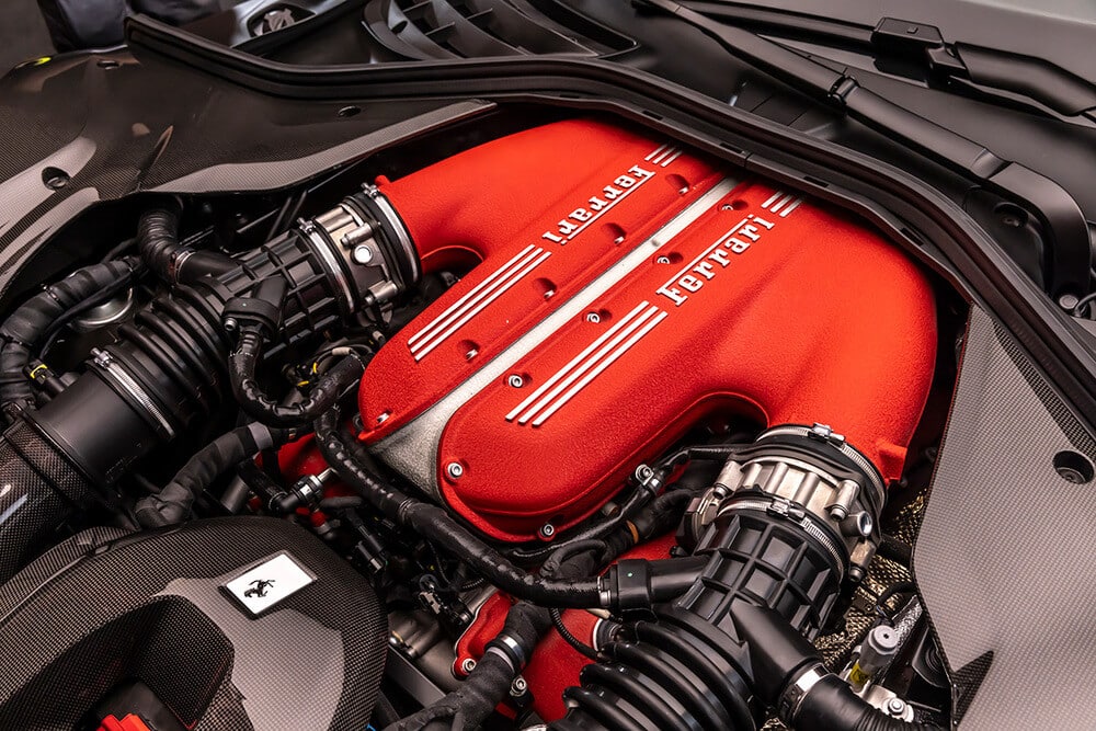La imagen muestra el compartimento del motor de un Ferrari, destacando específicamente el motor V12. La cubierta del motor está pintada de un color rojo distintivo con el logotipo de Ferrari exhibido de manera prominente. Se pueden ver los componentes del motor, incluyendo el sistema de admisión de aire y varias mangueras y conectores. Esta imagen es interesante y relevante porque muestra la ingeniería de alto rendimiento y el diseño de un motor V12 de Ferrari, conocido por su potencia y prestigio en el mundo automotriz.