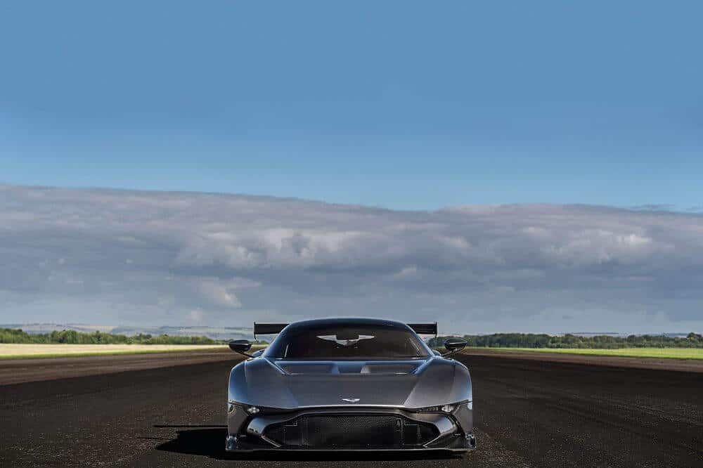 La imagen muestra un Aston Martin Vulcan de alto rendimiento posicionado en una amplia pista de aterrizaje vacía. El coche está orientado directamente hacia la cámara, lo que enfatiza su diseño aerodinámico y su agresiva fascia frontal. El fondo presenta un cielo azul claro con algunas nubes y un horizonte distante de campos verdes, sugiriendo un entorno abierto y expansivo. Esta imagen es interesante y relevante porque destaca el diseño y el potencial de rendimiento del coche, probablemente atrayendo a los entusiastas del automovilismo y mostrando las capacidades del vehículo en un entorno que sugiere velocidad y libertad.