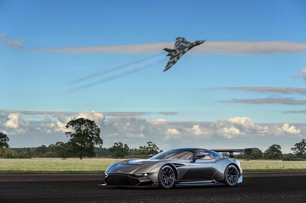 La imagen muestra un Aston Martin Vulcan negro estacionado en una pista de aterrizaje o carretera abierta. Sobre el coche, un avión militar, probablemente un bombardero o avión de reconocimiento, vuela en ángulo, dejando una estela de humo. El fondo presenta un cielo azul claro con algunas nubes y un campo de hierba con algunos árboles. La yuxtaposición del avanzado coche deportivo y el avión militar hace que la imagen sea interesante y visualmente impactante, destacando temas de velocidad, tecnología y excelencia en ingeniería.
