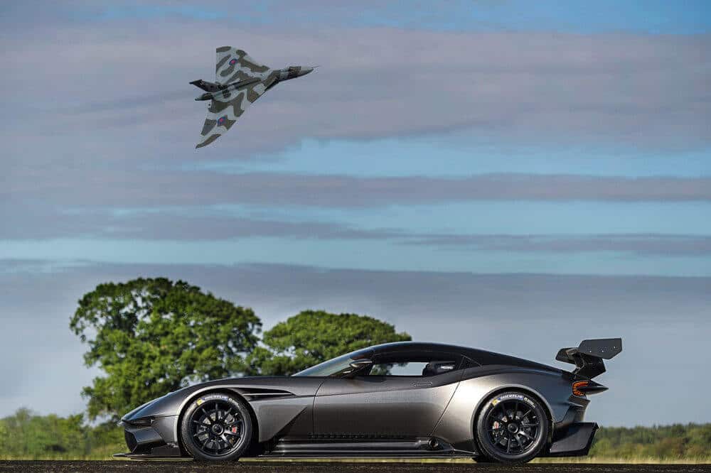La imagen muestra un Aston Martin Vulcan de alto rendimiento estacionado en una carretera con un gran avión militar camuflado volando por encima. El coche tiene un diseño aerodinámico con un gran alerón trasero, ruedas negras y neumáticos Michelin. El avión parece ser un jet de ala delta, posiblemente un bombardero o avión de reconocimiento, con patrones de camuflaje distintivos y escarapelas en sus alas. El fondo incluye un cielo despejado con algunas nubes y árboles verdes, sugiriendo un área rural o abierta. Esta imagen es interesante porque yuxtapone la ingeniería avanzada y el diseño estético tanto del coche como del avión, destacando la destreza tecnológica de ambos.