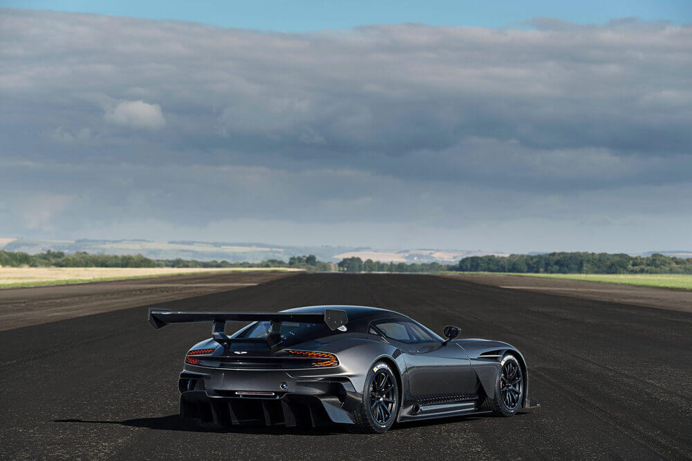 La imagen muestra un Aston Martin Vulcan de alto rendimiento en una pista de aterrizaje vacía. El coche está orientado con su parte trasera hacia el espectador, destacando su diseño aerodinámico, su gran alerón trasero y su estilo agresivo. El fondo presenta un paisaje abierto con campos y árboles distantes bajo un cielo nublado. Esta imagen es interesante y relevante porque resalta el diseño y las capacidades de rendimiento del coche, sugiriendo que está construido para la velocidad y la precisión en una pista o pista de aterrizaje.