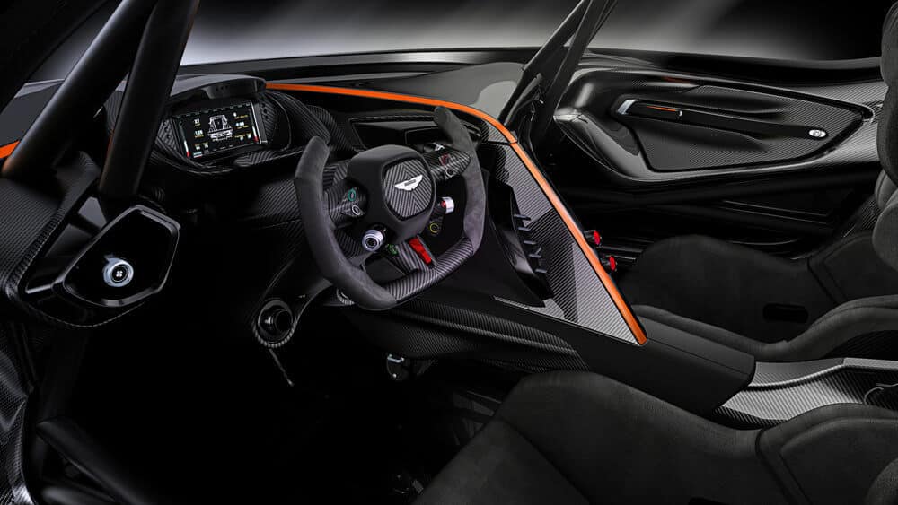 La imagen muestra el interior de un Aston Martin Vulcan, un coche deportivo de alto rendimiento. El tablero y el volante están hechos de fibra de carbono, y el volante tiene un diseño único con múltiples botones e interruptores, indicando funcionalidades avanzadas. El tablero incluye una pantalla digital que muestra varios indicadores de rendimiento. Los asientos son de estilo cubo, diseñados para la conducción a alta velocidad, y el diseño general enfatiza materiales ligeros y aerodinámicos. Esta imagen es interesante y relevante porque muestra la tecnología avanzada y los elementos de diseño utilizados en los vehículos modernos de alto rendimiento.
