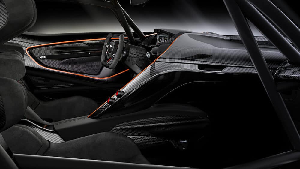 La imagen muestra el interior de un Aston Martin Vulcan, un coche deportivo de alto rendimiento. El diseño se centra en el lado del conductor, con un uso extensivo de materiales de fibra de carbono, lo que le da una apariencia elegante y moderna. El tablero y la consola central son aerodinámicos, con una pantalla digital detrás del volante. El volante tiene un diseño deportivo con múltiples controles. Los asientos están contorneados para brindar soporte, probablemente hechos de una combinación de fibra de carbono y Alcantara o materiales similares. Los acentos naranjas resaltan las líneas de diseño y añaden un toque de contraste de color. Esta imagen es interesante y relevante ya que muestra el diseño automotriz avanzado y los materiales utilizados en coches deportivos de alta gama, enfatizando el rendimiento y la estética.