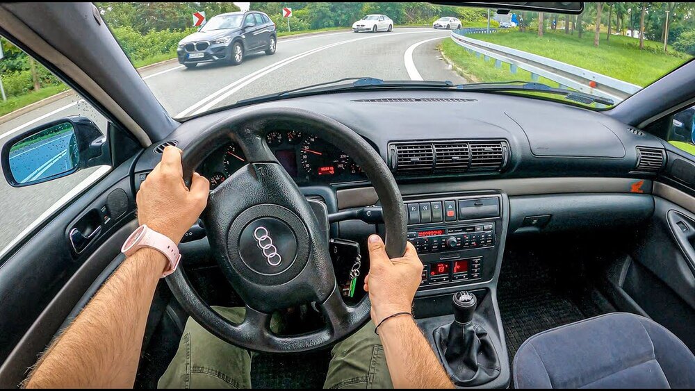 La imagen muestra la vista interior de un Audi A4 B5 desde la perspectiva del conductor. El conductor está sujetando el volante con ambas manos, y el coche está en una carretera curva con otros vehículos visibles a través del parabrisas. El salpicadero y la consola central del coche son visibles, con varios controles y pantallas. La imagen es interesante ya que proporciona una vista detallada de la experiencia de conducción y la disposición del interior del coche, lo cual puede ser relevante para los entusiastas de los coches o aquellos interesados en el modelo Audi A4 B5.