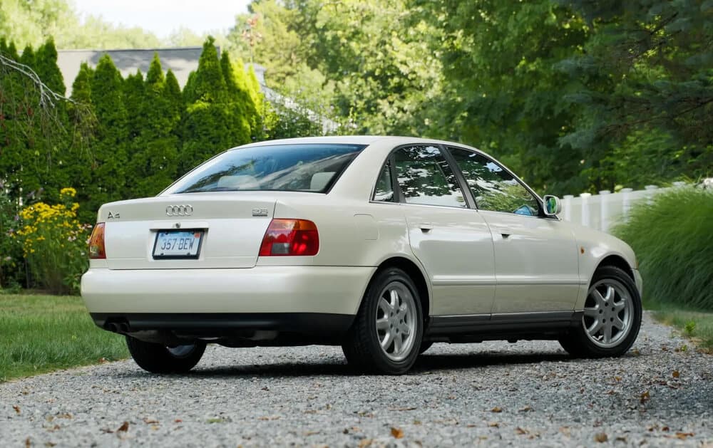 La imagen muestra la vista trasera y lateral de un Audi A4 B5 blanco estacionado en un camino de grava. El coche tiene una matrícula con el número '357-BEW'. El vehículo está rodeado por un jardín exuberante con árboles y arbustos en el fondo. El Audi A4 es un modelo bien conocido, y la imagen destaca su diseño y estado, lo cual podría ser de interés para los entusiastas de los coches o posibles compradores.