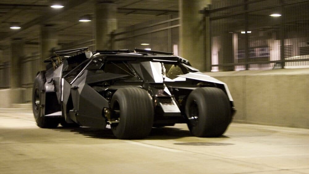 La imagen muestra el Batmóvil Tumbler, conocido por su diseño robusto y blindado, popularizado en las películas de Batman dirigidas por Christopher Nolan. El vehículo tiene grandes neumáticos gruesos y un exterior angular de color negro mate, lo que le da una apariencia intimidante y futurista. Este diseño incluye varios componentes mecánicos y detalles que sugieren un alto rendimiento y durabilidad, haciendo del Tumbler un vehículo icónico y fascinante tanto en la ficción como en la realidad.