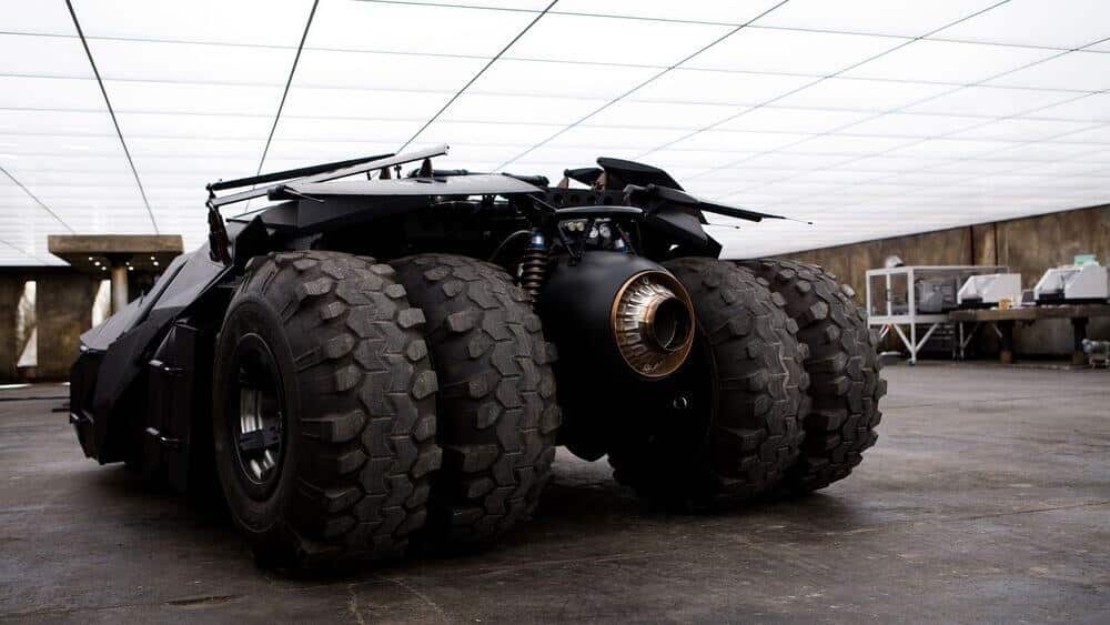 La imagen muestra una vista trasera del Batmóvil Tumbler, un vehículo ficticio de la franquicia de Batman. El Tumbler se caracteriza por su diseño robusto y militar, con grandes neumáticos todoterreno y un prominente escape de motor a reacción en la parte trasera. El vehículo está situado en lo que parece ser un garaje o taller de alta tecnología, con varios equipos y bancos de trabajo visibles en el fondo. Esta imagen es interesante y relevante debido al estatus icónico del Batmóvil en la cultura popular, especialmente en su representación en la trilogía de películas "The Dark Knight".