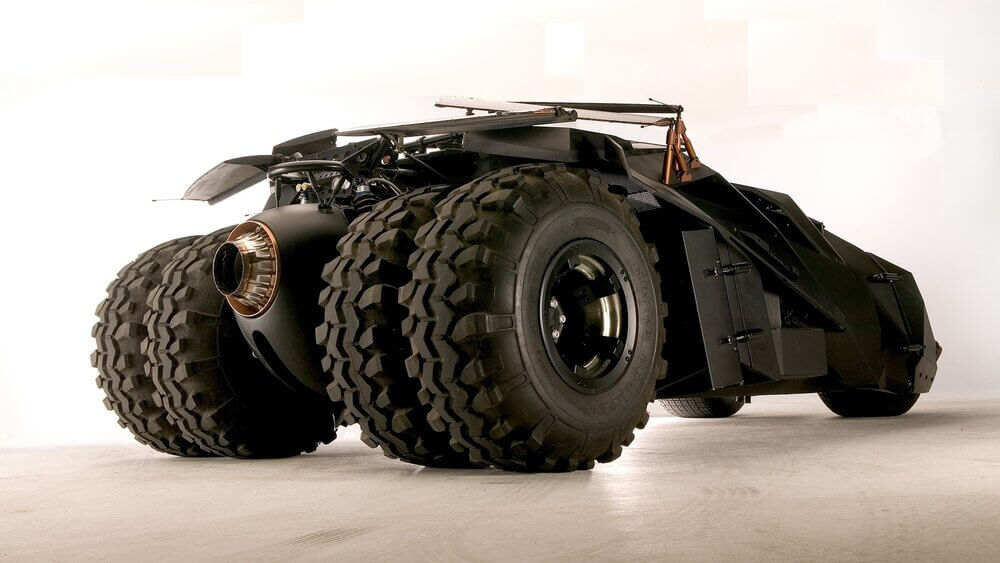 La imagen muestra el Batmóvil Tumbler, conocido por su diseño robusto y blindado, popularizado en las películas de Batman dirigidas por Christopher Nolan. El vehículo tiene grandes neumáticos gruesos y un exterior angular de color negro mate, lo que le da una apariencia intimidante y futurista. Este diseño incluye varios componentes mecánicos y detalles que sugieren un alto rendimiento y durabilidad, haciendo del Tumbler un vehículo icónico y fascinante tanto en la ficción como en la realidad.