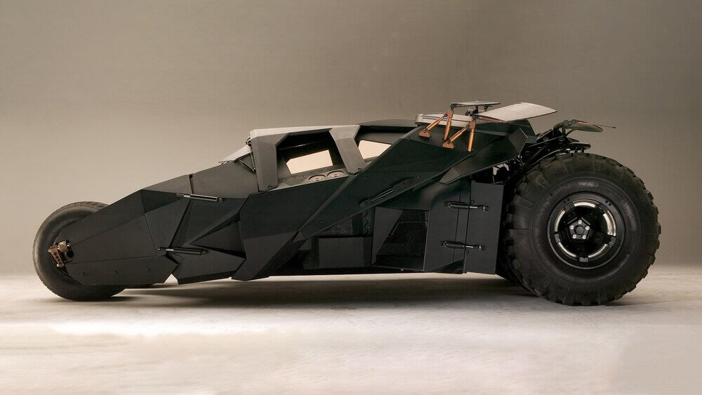 La imagen muestra una vista lateral de un vehículo conocido como el Batmóvil. Este Batmóvil en particular tiene un diseño futurista y blindado, con una carrocería angular y elegante, grandes neumáticos traseros y un neumático delantero más pequeño. El vehículo es predominantemente negro y tiene una apariencia robusta y táctica, característica del Batmóvil utilizado por el superhéroe ficticio Batman. Esta imagen es interesante y relevante porque el Batmóvil es un elemento icónico de la franquicia de Batman, conocido por su tecnología avanzada y diseño único.