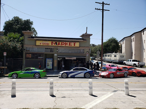 Gracias por la corrección. La imagen muestra un mercado de alimentos, un lugar notable de la franquicia de películas "Fast & Furious", específicamente la tienda propiedad del personaje Dominic Toretto. Frente al mercado, hay cuatro coches deportivos personalizados y de colores brillantes estacionados en fila. Los coches son un Mitsubishi Eclipse verde, un Mazda RX-7 rojo, un Acura NSX rojo y un BMW M3 azul y plateado, pintado como el M3 GTR de "Most Wanted". Hay varias personas cerca de la entrada del mercado, y parece ser un día soleado. El mercado tiene un cartel que dice "FOOD MARKET" en letras grandes. La imagen es interesante y relevante porque representa un lugar famoso de una serie de películas popular, y la presencia de los coches icónicos añade a su importancia para los fanáticos de la franquicia.