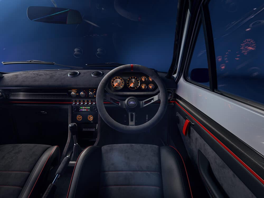 La imagen muestra el interior de un Ford Escort MK1 de Boreham Motorworks. El volante está en el centro, con el logotipo de Ford en el medio. El tablero presenta varios indicadores con retroiluminación naranja, incluyendo un velocímetro y un tacómetro. Debajo del tablero, hay controles adicionales y botones, así como una palanca de cambios y un freno de mano. El diseño interior incluye acentos en negro y rojo, con una combinación de materiales de cuero y gamuza. El aspecto general es deportivo y moderno, sugiriendo que este podría ser un vehículo de alto rendimiento o personalizado.