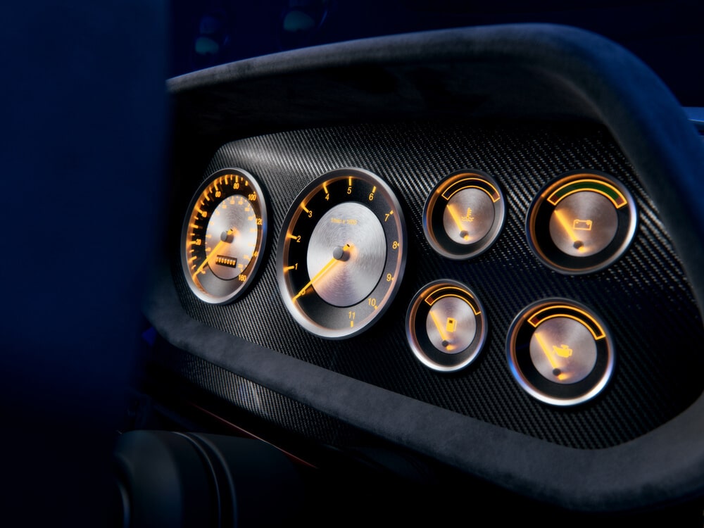 La imagen muestra el tablero de un Ford Escort MK1 de Boreham Motorworks. El tablero presenta varios indicadores analógicos con retroiluminación naranja. Los indicadores incluyen un velocímetro, un tacómetro y varios medidores más pequeños para el nivel de combustible, la temperatura del motor, el voltaje de la batería y la presión del aceite. El diseño es elegante y moderno, con un fondo de fibra de carbono y acentos metálicos, lo que lo hace visualmente atractivo e indicativo de un vehículo de alto rendimiento o de lujo.