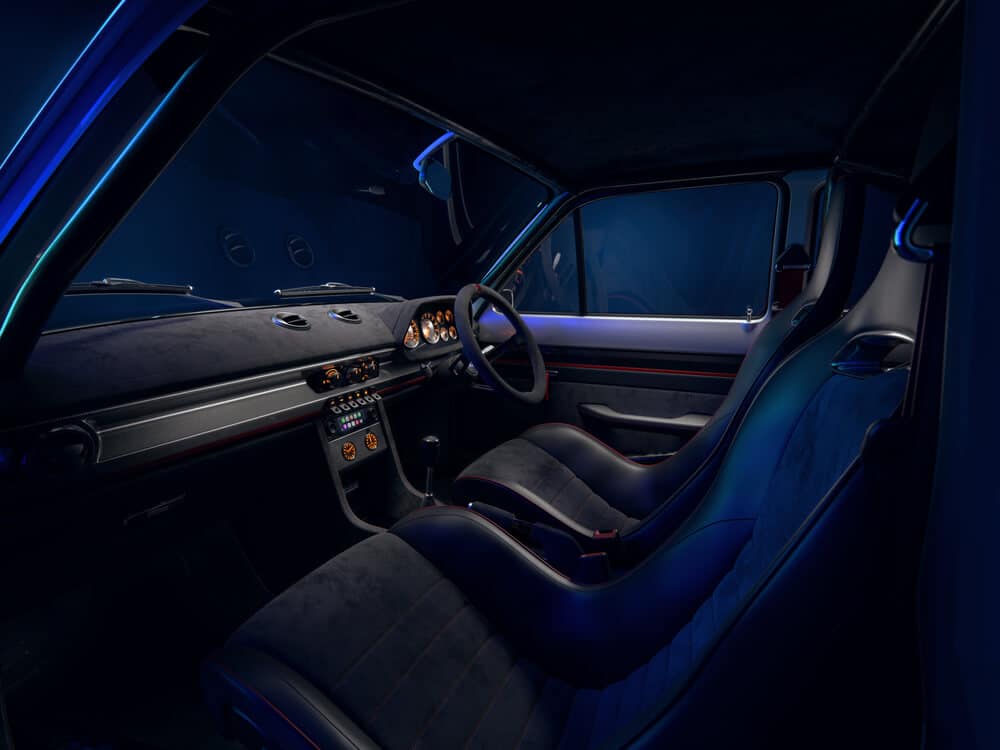 La imagen muestra el interior de un Ford Escort MK1 de Boreham Motorworks. El enfoque está en los asientos delanteros, el tablero y el volante. El diseño interior es moderno y elegante, con un esquema de colores oscuros que incluye tonos negros y azul oscuro. Los asientos son deportivos y contorneados, probablemente hechos de cuero o un material similar, con costuras rojas. El tablero incluye varios indicadores y controles, lo que indica que es un vehículo de alto rendimiento. La iluminación dentro del coche tiene un tono azul, añadiendo a la atmósfera moderna y sofisticada.