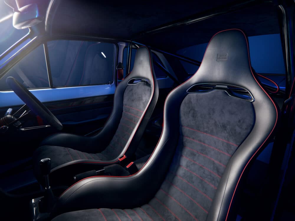 La imagen muestra el interior de un Ford Escort MK1 de Boreham Motorworks. El enfoque está en los asientos delanteros, el tablero y el volante. El diseño interior es moderno y elegante, con un esquema de colores oscuros que incluye tonos negros y azul oscuro. Los asientos son deportivos y contorneados, probablemente hechos de cuero o un material similar, con costuras rojas. El tablero incluye varios indicadores y controles, lo que indica que es un vehículo de alto rendimiento. La iluminación dentro del coche tiene un tono azul, añadiendo a la atmósfera moderna y sofisticada.