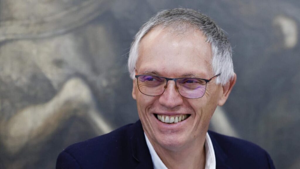 La imagen muestra a Carlos Tavares, el CEO de Stellantis, sonriendo. Está vestido con un traje oscuro y una camisa blanca, y el fondo parece ser una pintura o mural desenfocado con formas y colores indistintos, principalmente en tonos de gris y marrón. La imagen es interesante porque captura a Tavares en un momento de alegría o satisfacción, lo cual puede ser relevante en el contexto de una presentación, entrevista o aparición pública.
