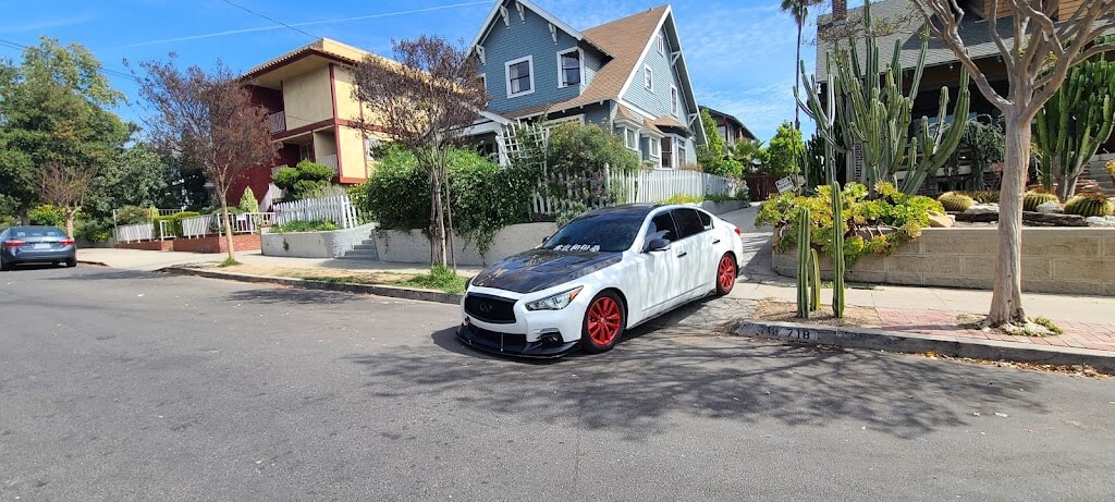 La imagen muestra una calle residencial con varias casas y un coche blanco estacionado al lado de la carretera. El coche tiene llantas rojas y un capó negro, y hay texto en el parabrisas. Las casas en el fondo son de diferentes estilos arquitectónicos, con una casa azul y otra roja y amarilla. Hay varias plantas y árboles a lo largo de la acera, incluyendo un gran cactus. La escena parece estar en un vecindario suburbano. Esta imagen es interesante porque captura un ambiente cotidiano y tranquilo, que contrasta con la acción y el drama típicos de las películas de "Fast & Furious". Es la casa de Toretto.