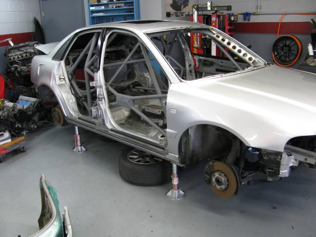 "La imagen muestra el chasis de un Audi A4 B5 en un taller. El coche está elevado sobre soportes, con las puertas quitadas y el interior desmantelado, revelando una estructura de jaula antivuelco en su interior. Las ruedas y varios otros componentes han sido retirados, lo que indica que el coche está siendo sometido a modificaciones o reparaciones significativas. El taller está equipado con varias herramientas y equipos, incluyendo un polipasto de motor y piezas de repuesto, lo que sugiere un entorno profesional o de entusiastas enfocado en el trabajo automotriz. Esta imagen es interesante y relevante ya que muestra el proceso detallado de modificación o restauración de un coche, destacando la complejidad y minuciosidad involucradas en tales proyectos.