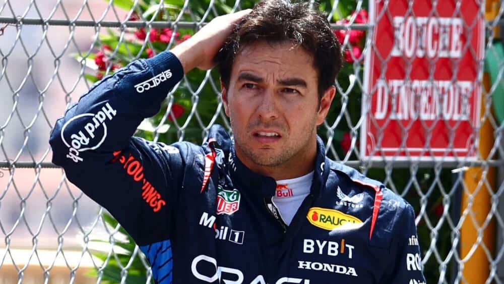 La imagen muestra a Checo Pérez, piloto de Fórmula 1, con un traje de carreras que lleva varios logotipos de patrocinadores, incluyendo Oracle, Bybit, Honda y Hard Rock. Checo está de pie frente a una valla de cadena con un cartel rojo que dice "BOUCHE D'INCENDIE." Tiene una mano en la cabeza, lo que sugiere un momento de preocupación o reflexión, posiblemente relacionado con un evento de carreras.