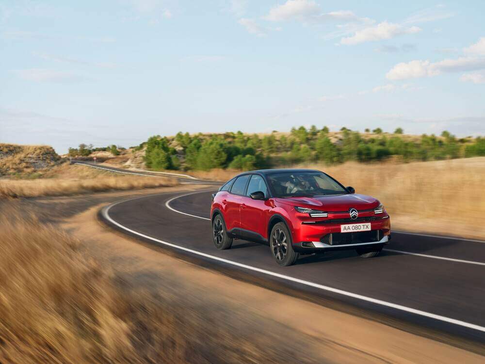 "Imagen de un coche Citroën rojo conduciendo a alta velocidad por una carretera sinuosa a través de un paisaje seco y con hierba, con algunos árboles verdes al fondo. La matrícula del coche dice 'AA 080 TX'. La imagen captura el rendimiento dinámico y el diseño del vehículo en un entorno pintoresco, destacando las capacidades y el atractivo estético del coche.