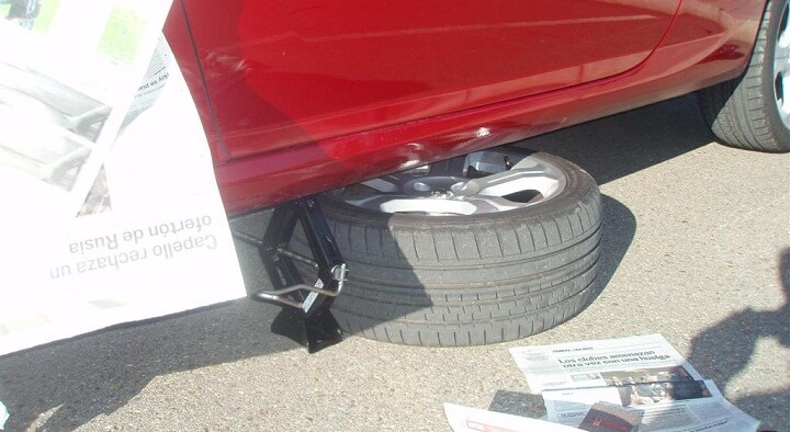 "Imagen de un coche rojo levantado con un gato. La rueda ha sido retirada y colocada debajo del coche como medida de seguridad. Hay periódicos esparcidos en el suelo alrededor del coche, posiblemente para mantener el área limpia o proporcionar algo de amortiguación.