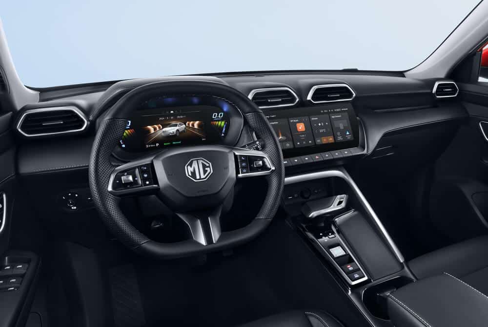 La imagen muestra el interior de un vehículo MG ZS Hybrid+, específicamente enfocándose en el área del conductor. El volante presenta el logotipo de MG prominentemente en el centro. Detrás del volante hay un cuadro de instrumentos digital que muestra información variada como la velocidad, la posición de la marcha y una representación visual del coche. A la derecha del volante hay un sistema de infoentretenimiento con pantalla táctil grande integrado en el tablero, mostrando varios controles y configuraciones del vehículo. El diseño del tablero es moderno y elegante, con rejillas de ventilación y botones de control dispuestos de manera ordenada. La consola central incluye un selector de marchas y controles adicionales. Esta imagen es interesante y relevante ya que destaca la tecnología avanzada y las características de diseño moderno del interior del MG ZS Hybrid+.
