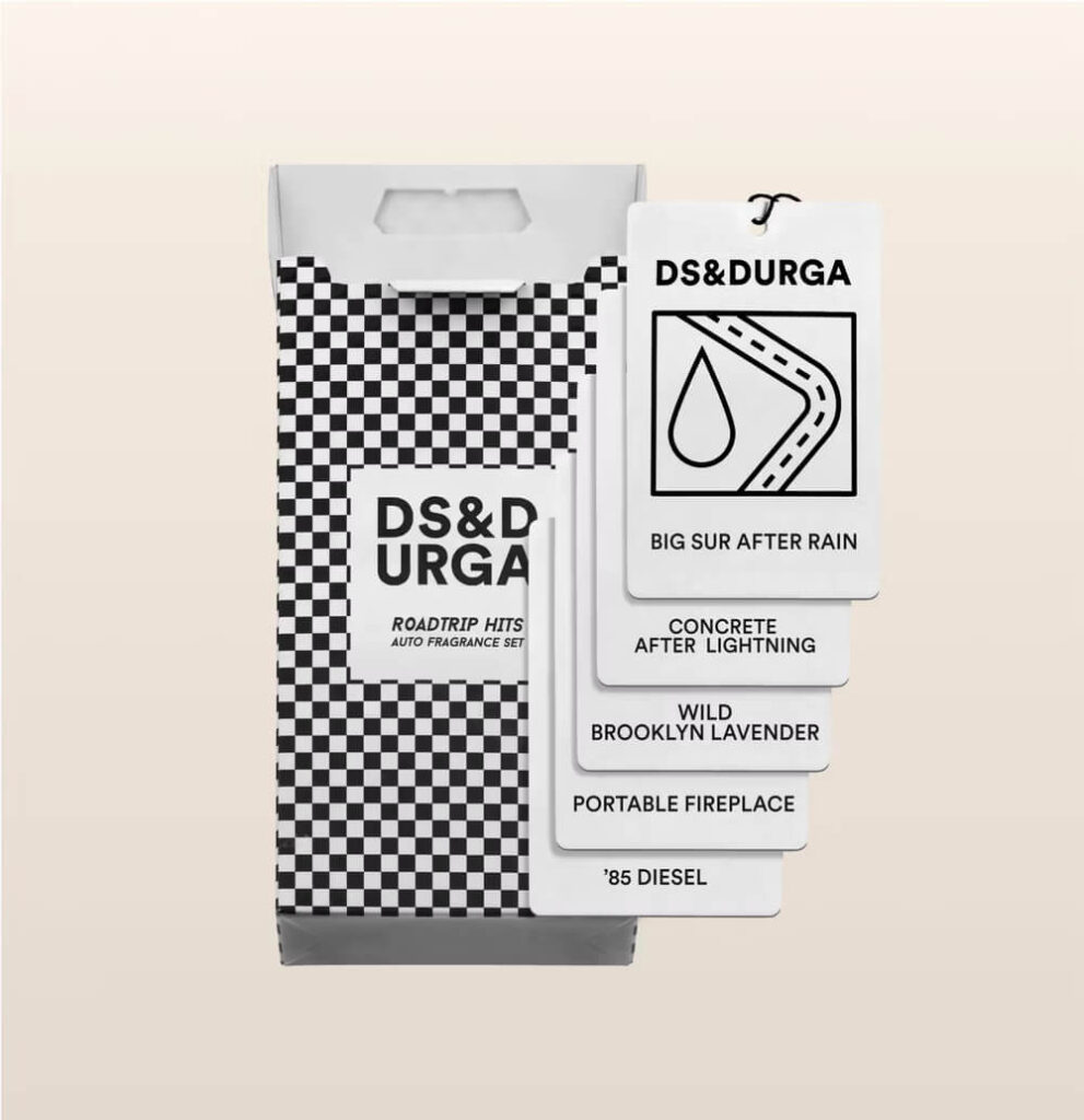 La imagen muestra el empaque de un set de fragancias para auto de DS & DURGA llamado "Roadtrip Hits Auto Fragrance Set." El empaque tiene un diseño a cuadros en blanco y negro. Se muestran cinco tarjetas de fragancia, cada una con un nombre de aroma diferente. Los aromas listados son:

Big Sur After Rain

Concrete After Lightning

Wild Brooklyn Lavender

Portable Fireplace

'85 Diesel

Esta imagen es interesante porque presenta un conjunto único de fragancias para autos con nombres creativos y evocadores, sugiriendo una experiencia sensorial relacionada con lugares y momentos específicos.