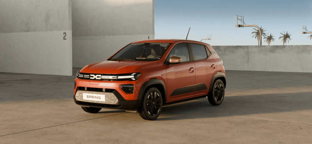 Dacia Spring 2024 en color naranja cobrizo, fotografiado en un espacio minimalista de hormigón con palmeras al fondo. El coche, un SUV compacto eléctrico, muestra su nuevo diseño frontal con la parrilla iluminada en forma de 'DC', faros LED afilados, y detalles en negro como las llantas y las molduras laterales. Está fotografiado en ángulo tres cuartos delantero, mostrando su silueta elevada y robusta característica de un crossover urbano. En el fondo se pueden ver canastas de baloncesto y algunas palmeras contra un cielo azul claro.