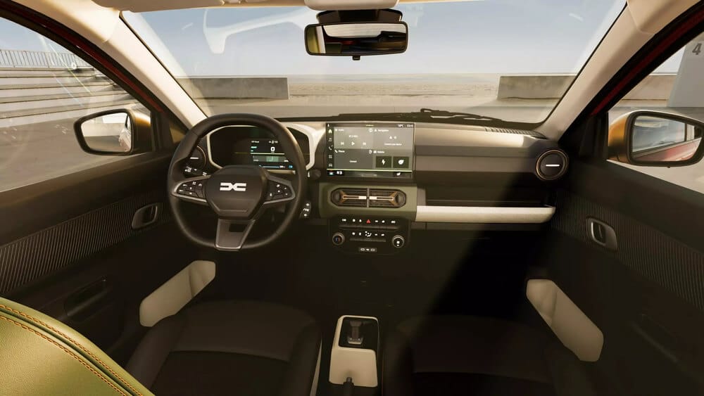 Interior del Dacia Spring 2024 fotografiado desde el asiento trasero, mostrando el puesto de conducción completo. Destaca una pantalla digital como cuadro de instrumentos, una pantalla central táctil para el sistema multimedia, y un volante con el nuevo logo 'DC' de Dacia. El salpicadero presenta un diseño minimalista con una franja decorativa horizontal en color claro, controles de climatización físicos bajo la pantalla central, y tapicería en tonos oscuros con detalles en beige. La consola central es compacta y cuenta con el selector de marchas. El interior combina plásticos texturizados con inserciones en tonos claros para dar un aspecto más moderno.