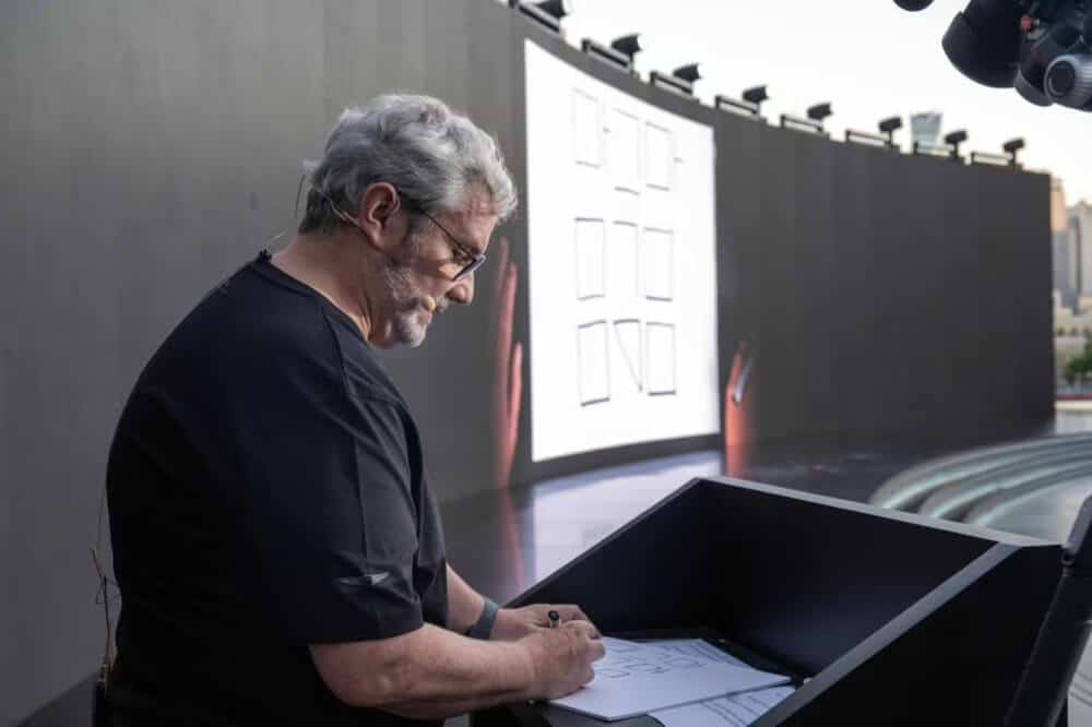 Luc Donckerwolke dibuja el logotipo de Genesis Magma Racing en un podio o escritorio. Lleva una camisa negra y tiene un micrófono de auriculares. En el fondo, hay una gran pantalla curva que muestra una serie de rectángulos y líneas, que parecen ser diagramas o bocetos.